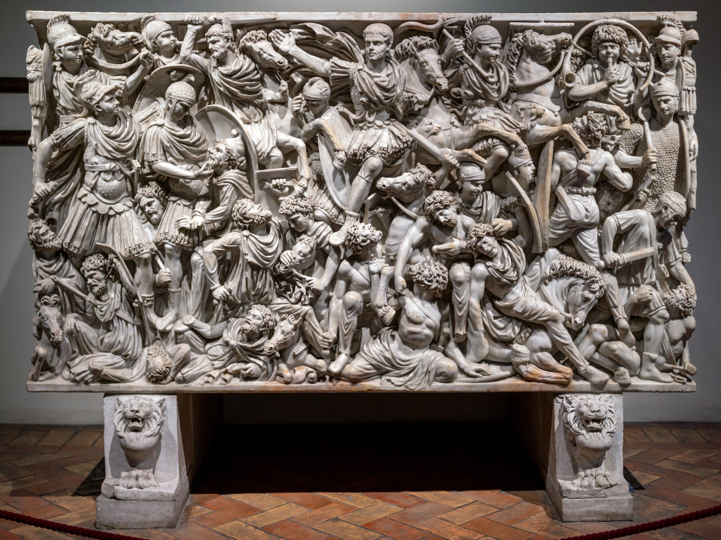A detailed photograph of an ancient Roman marble sarcophagus featuring a high-relief battle scene with numerous warriors, horses, and weapons intricately carved in dynamic poses. The sarcophagus rests on two lion-headed supports and showcases craftsmanship through expressive figures, overlapping elements, and a sense of movement capturing a dramatic combat moment.