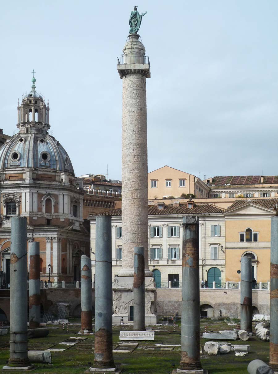 Photograph of Trajan's Column, a Roman triumphal column in Rome, Italy, surrounded by ancient ruins and historic buildings. The tall, cylindrical column features a spiral bas-relief and is topped with a statue, set against a cloudy sky.