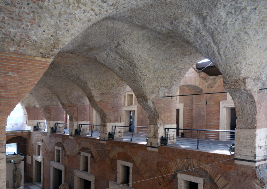 Photograph of an ancient Roman architectural interior featuring large stone arches and brick walls. The space includes a mezzanine level with metal railings and doorways framed by stone, showcasing historical construction techniques and structural design.
