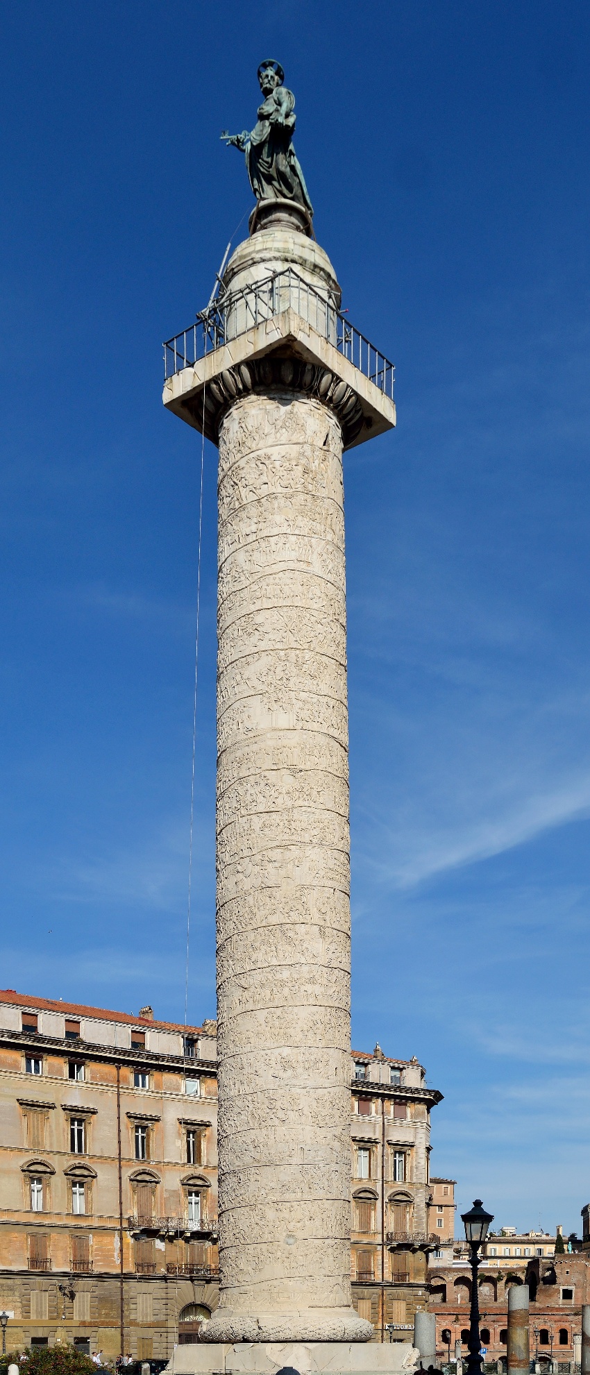 Photograph of Trajan's Column, a Roman triumphal column in Rome, Italy, featuring a spiral bas-relief that commemorates Emperor Trajan's victory in the Dacian Wars. The column is topped with a statue and stands prominently against a clear blue sky, with historic buildings visible at its base.