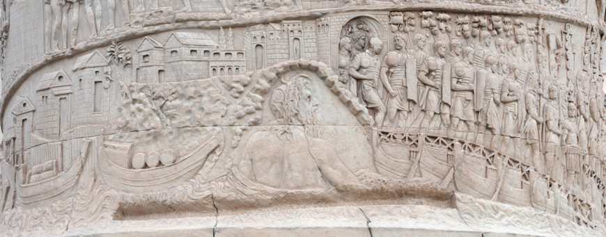 Bas-relief sculpture depicting an ancient Roman military scene with soldiers, boats, and architectural structures. Detailed carvings show soldiers in formation, a bridge, and various elements of a cityscape, illustrating a historical campaign.