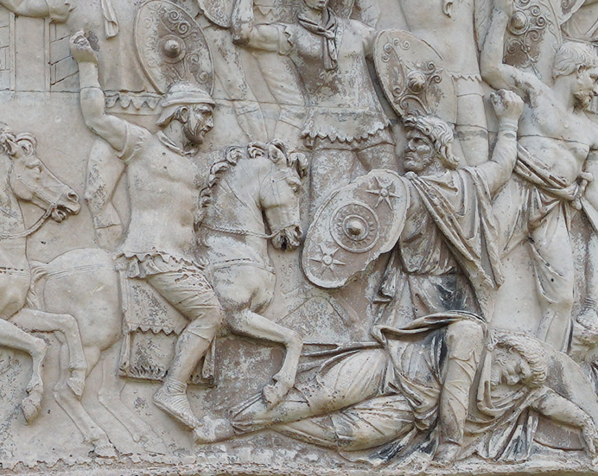A detailed stone relief sculpture depicting a battle scene with armored soldiers on horseback and foot soldiers engaged in combat. Key elements include raised shields, helmets, and dynamic poses conveying movement and conflict, with intricate carvings highlighting armor and weaponry.