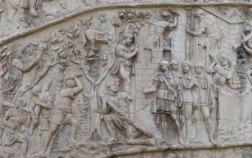 Bas-relief sculpture depicting a historical scene with multiple human figures engaged in various activities, including building, climbing, gesturing, and conversing. Carved in stone with detailed clothing and architectural elements, the artwork highlights interaction and movement within an ancient setting.