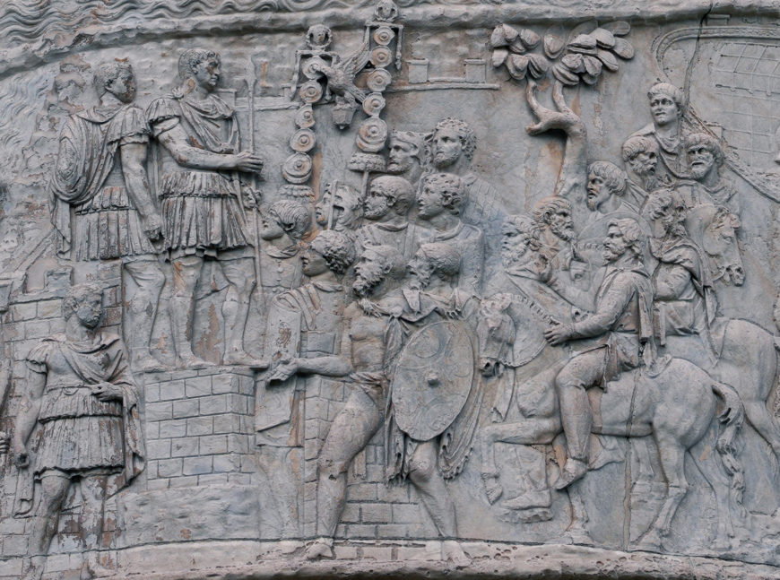 Bas-relief sculpture depicting a historical Roman scene with soldiers and figures in classical attire, some holding shields and standards. The composition features a group on horseback and others standing, highlighting military and ceremonial elements carved in stone with detailed facial expressions and attire.