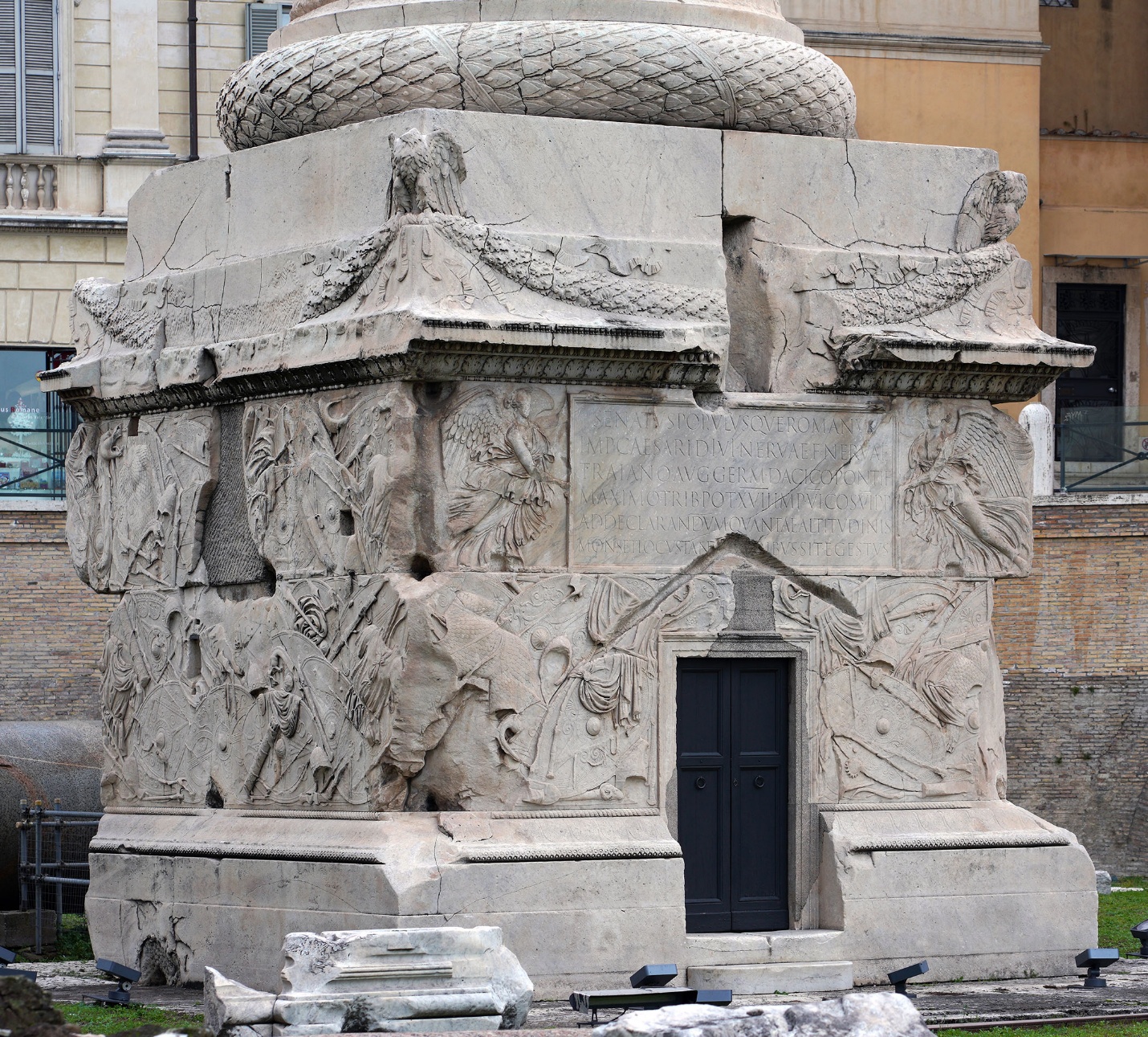 Photograph of Base (detail), Column of Trajan Roman stone monument featuring intricate bas-relief carvings of figures, animals, and decorative motifs on all visible sides. The structure includes a central doorway and a rectangular inscription panel,