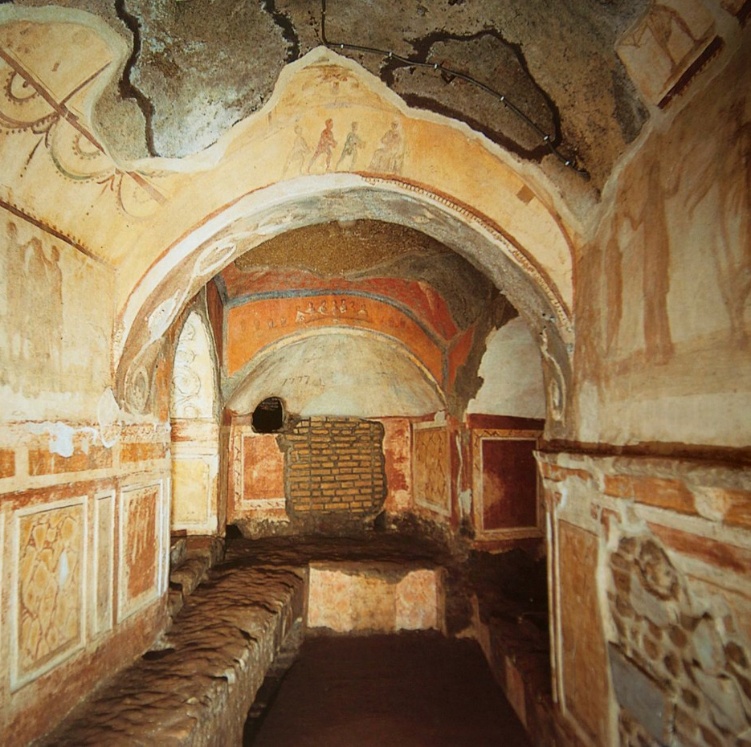 Photograph of an ancient underground catacomb with arched ceilings and walls adorned with faded frescoes and murals in earthy tones. The space features a central stone bench, a bricked-up niche, and detailed decorative panels.