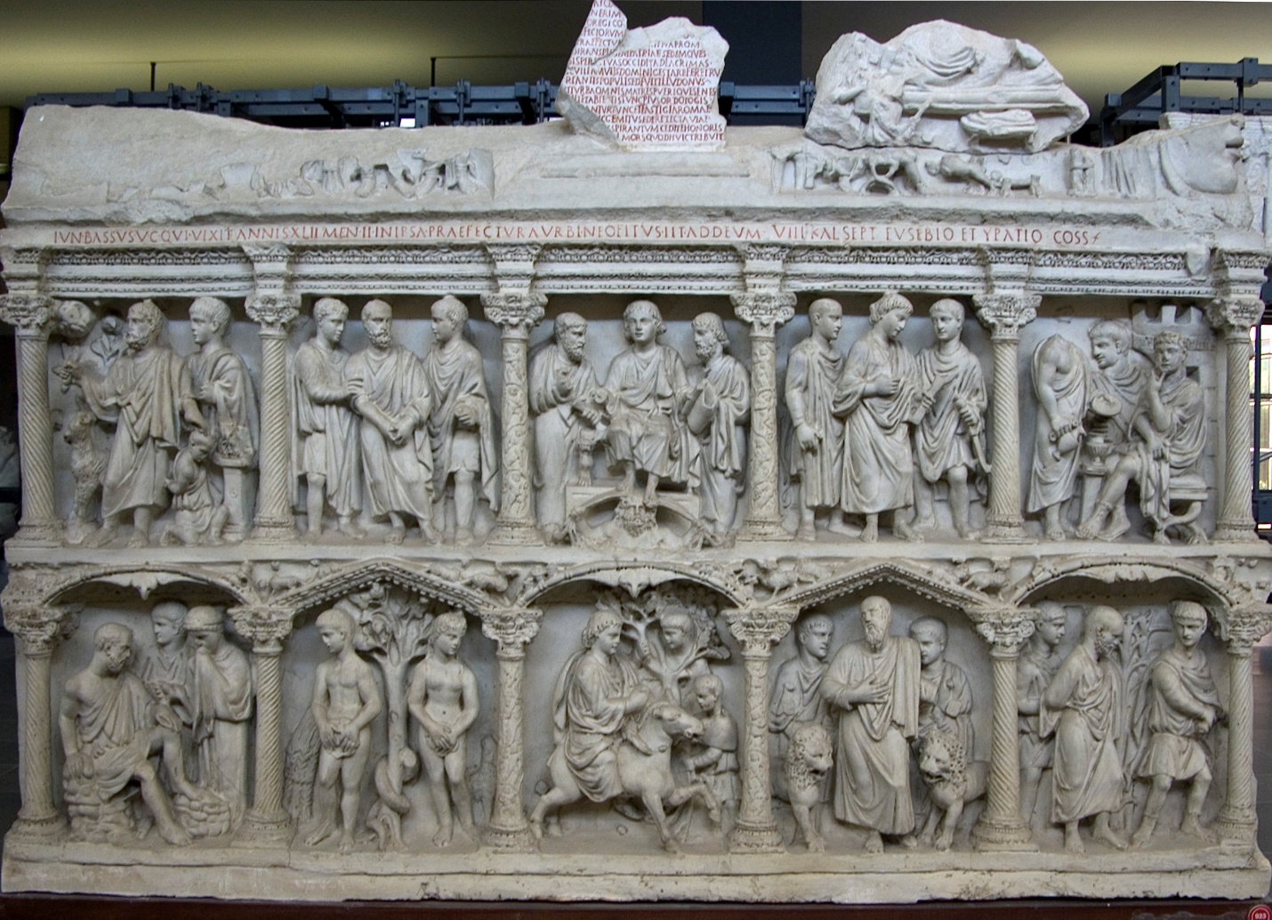 A photograph of an ancient Roman sarcophagus featuring detailed relief sculptures arranged in two horizontal registers. The upper register shows a series of standing figures in togas engaged in various interactions, while the lower register depicts scenes with both standing and seated figures, including a central figure on horseback, all framed by ornate columns and arches.