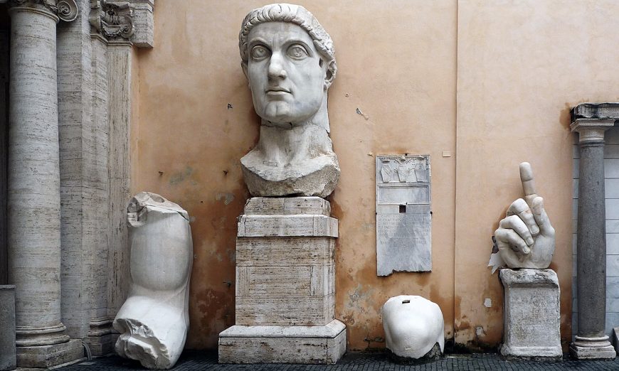 Photograph of ancient marble sculptures displayed against a beige wall, featuring a large head bust on a pedestal, a detached arm, a hand with an extended index finger, and a smaller rounded fragment. The sculptures are arranged on a cobblestone ground between stone columns, highlighting classical Roman art and architectural elements.