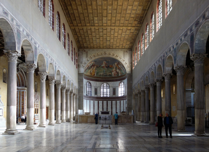 Photograph of a large, historic basilica interior featuring two rows of tall, ornate columns with arches supporting high walls and a coffered ceiling. The far end displays a semicircular apse with religious frescoes above arched windows, and a few people stand on the marble floor, emphasizing the vast, open space.
