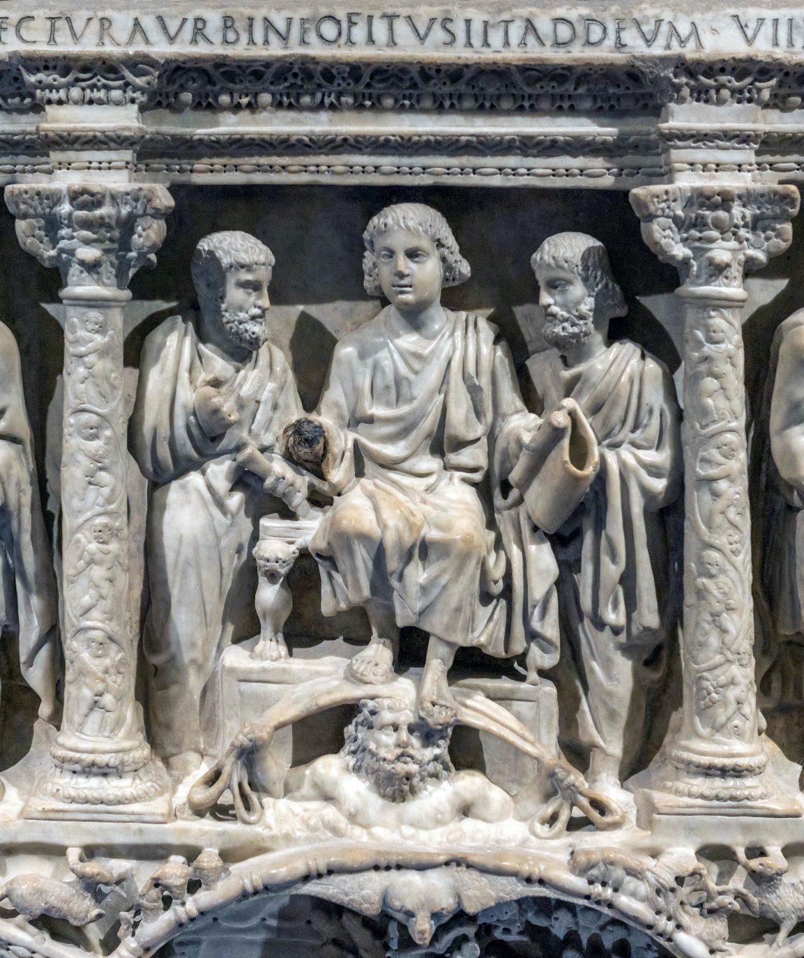 A detailed photograph of an ancient Roman marble relief sculpture depicting two robed figures standing behind a seated central figure, who is elevated on a platform supported by a muscular figure beneath. The relief features ornate columns on either side, Latin inscriptions above, and intricate carvings of animals and decorative elements along the base.