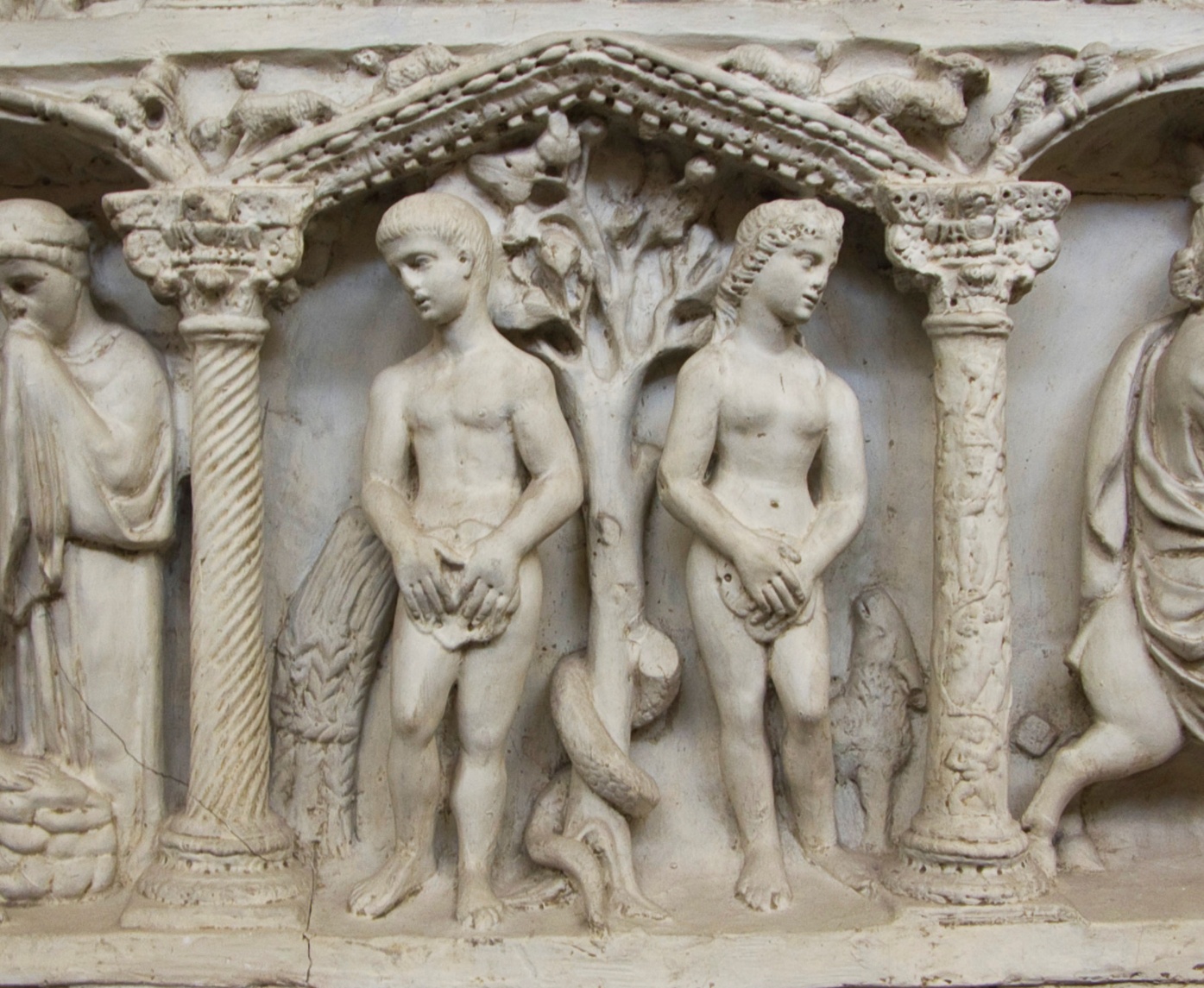 A detailed photograph of an ancient Roman marble relief sculpture depicting Adam and Eve in nude on either side of a tree wrapped with a serpent. The figures are flanked by two ornate columns.