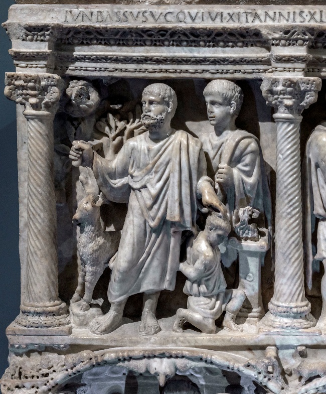 Marble relief sculpture depicting two robed male figures standing between two fluted columns with Corinthian capitals, accompanied by animals and a kneeling child. Latin inscription runs along top edge, highlighting Roman artistic and cultural themes.