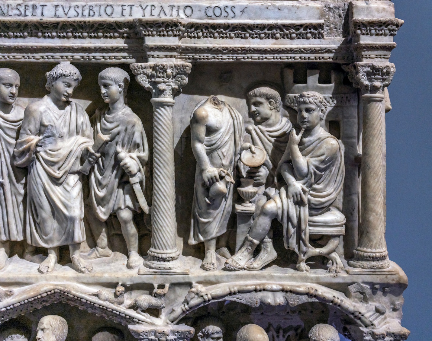 A photograph of an ancient marble relief sculpture depicting a group of robed figures engaged in a scene, framed by intricately carved columns and architectural elements. The relief features detailed drapery, a seated figure holding a scroll, and Latin inscriptions along the top edge, highlighting classical artistic style and historical context.