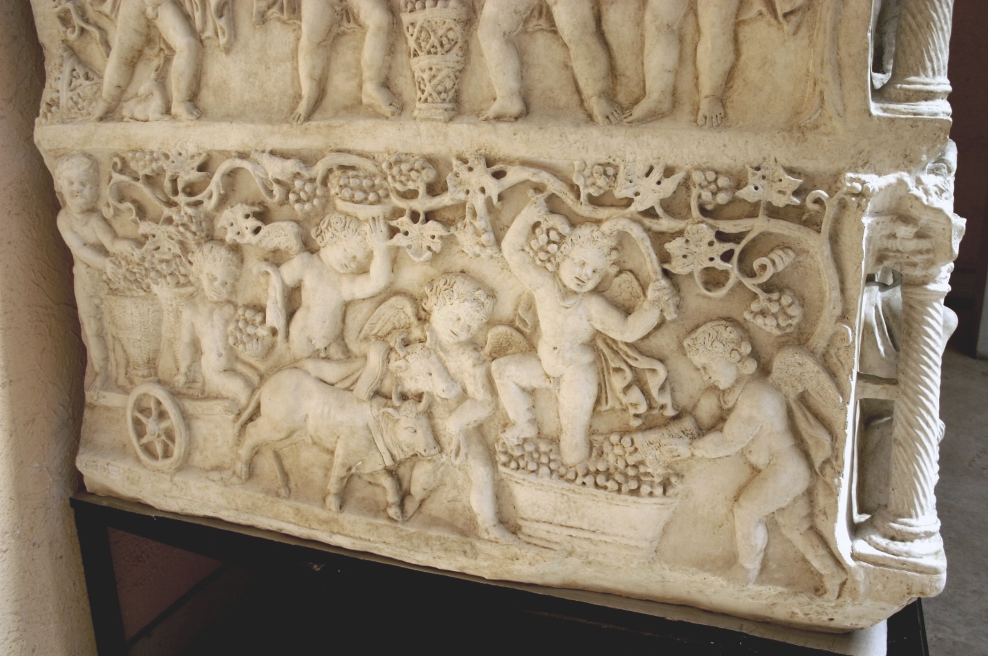 A photograph of a detailed stone relief sculpture depicting cherubic figures harvesting grapes and interacting with animals. The relief features intricate vine and grape motifs, with cherubs in dynamic poses, highlighting themes of abundance and pastoral life.