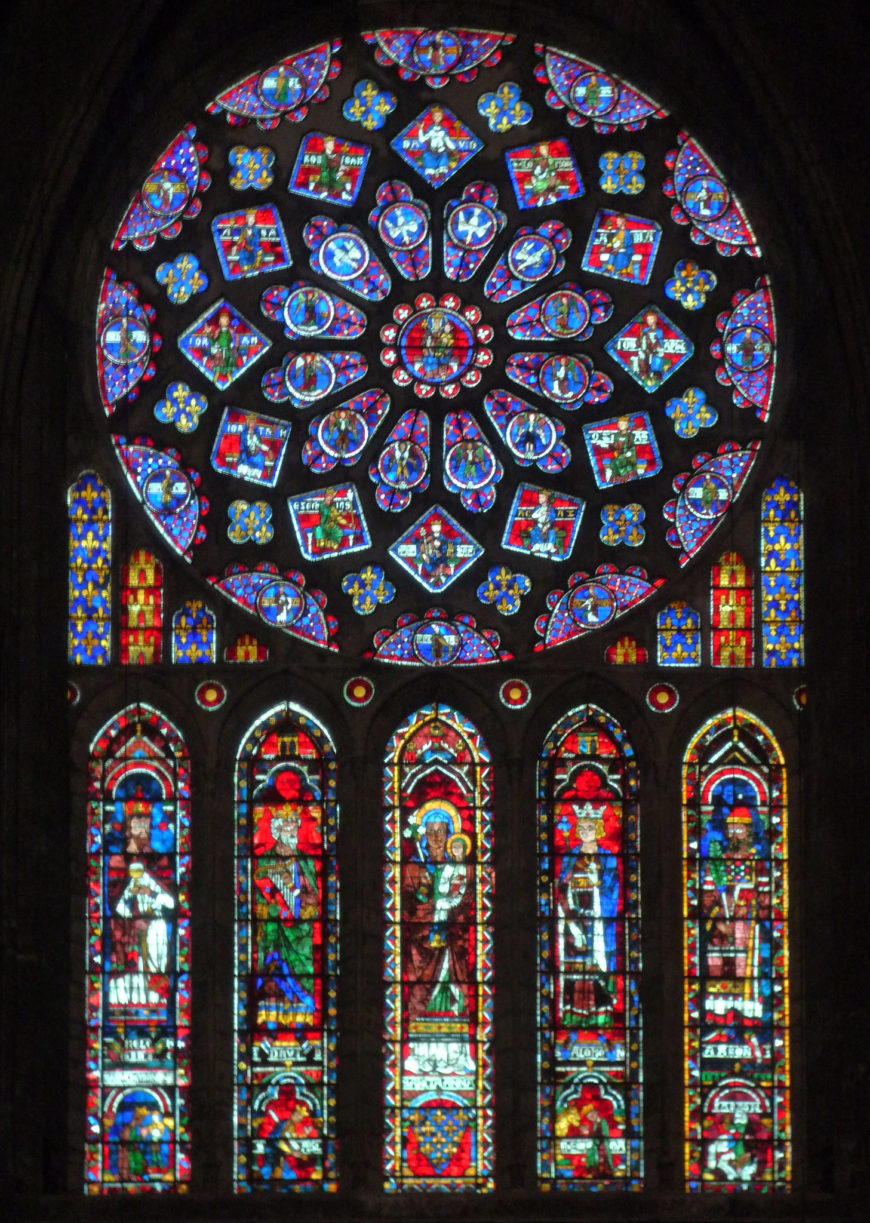 Stained glass window featuring a large circular rose design above five tall, narrow panels depicting religious figures in vibrant reds, blues, and greens. The intricate patterns and detailed imagery highlight traditional Gothic cathedral artistry and religious storytelling.