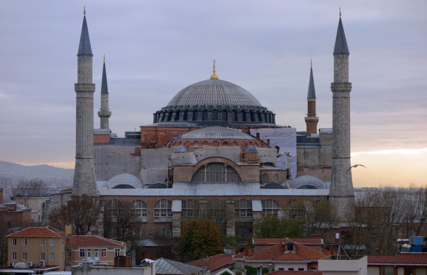 Photograph of Hagia Sophia in Istanbul during sunset, showcasing its large central dome and four tall minarets. The historic structure is surrounded by residential buildings and trees, with a cloudy sky in the background.