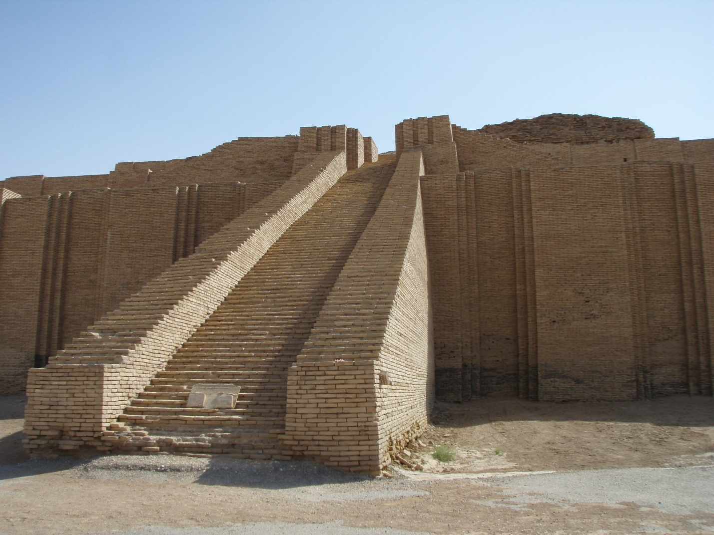 Photograph of an ancient stepped brick ziggurat with two wide staircases leading to the top platform, set against a clear blue sky. The structure features vertical brick patterns on its facade and shows signs of weathering.