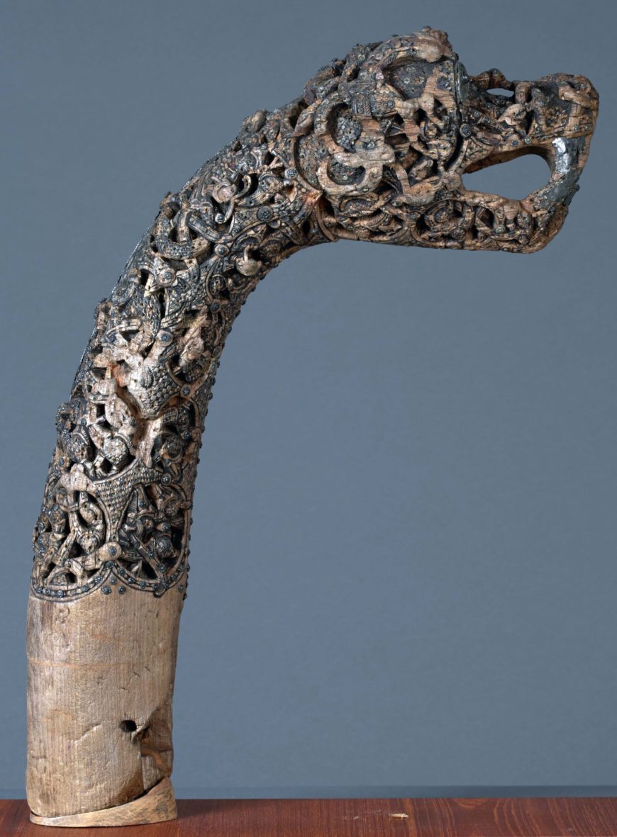 Photograph of an intricately carved wooden object shaped like a serpent or dragon head with an open mouth. The upper part features detailed openwork designs with intertwined patterns and figures, while the lower part is smooth and uncarved, resting on a wooden surface against a plain background.
