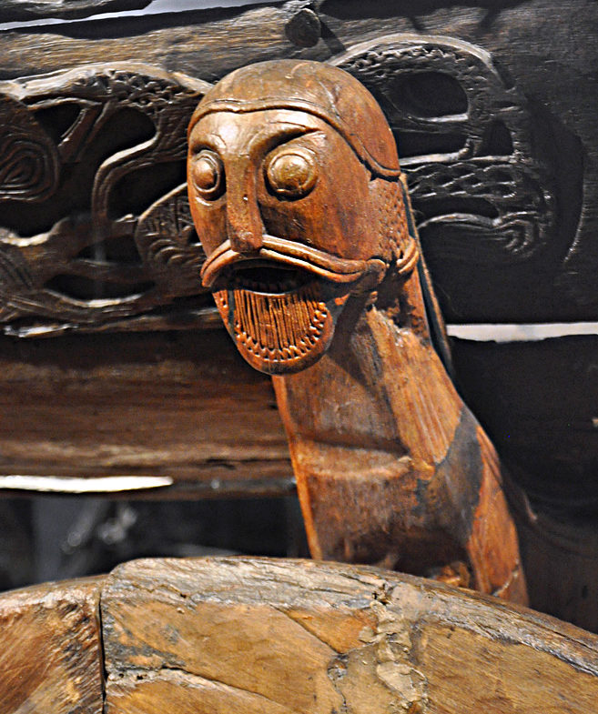 Photograph of a carved wooden figurehead featuring a stylized human face with large round eyes, a mustache, and an open mouth with vertical lines suggesting teeth. The figurehead is attached to a larger wooden structure with intricate carved patterns in the background, indicating traditional craftsmanship.