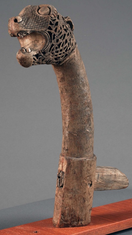Photograph of a carved wooden sculpture featuring a long, curved neck with a detailed animal head, possibly a dragon or serpent, at the top. The head showcases intricate lattice patterns and textured scales, mounted on a rectangular wooden base with a small protruding handle.