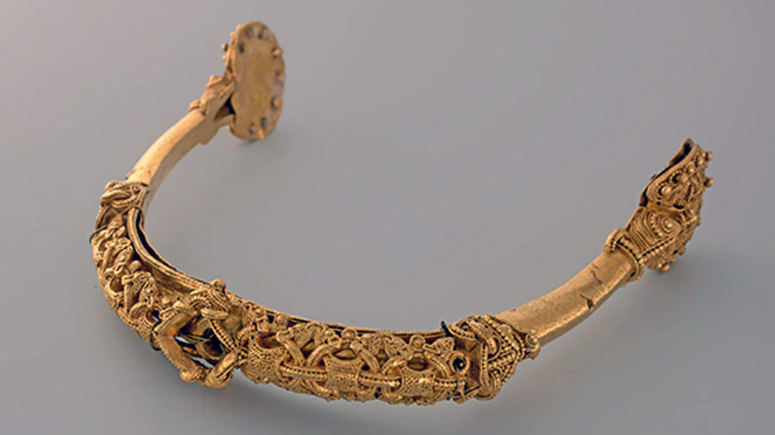 Photograph of an ornate gold spur featuring intricate filigree work and detailed patterns along its curved structure. The spur displays symmetrical designs with small decorative elements and a clasp mechanism at one end.