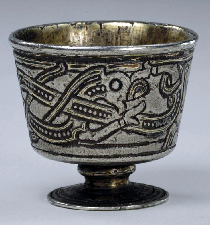 Photograph of an ancient metal goblet with intricate engraved designs featuring intertwined animal motifs and geometric patterns. The goblet has a wide rim, a slightly flared body, and a round pedestal base, showcasing craftsmanship and artistic style from a historical period.