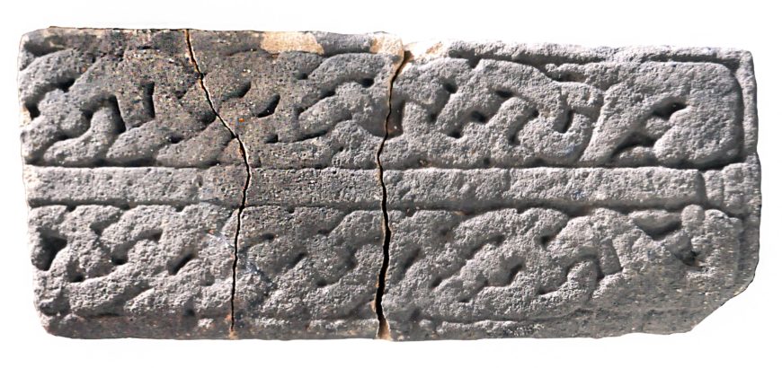 Photograph of an ancient stone tablet featuring intricate carved patterns arranged in two horizontal bands. The tablet shows signs of age with visible cracks and weathering, highlighting detailed interwoven designs.