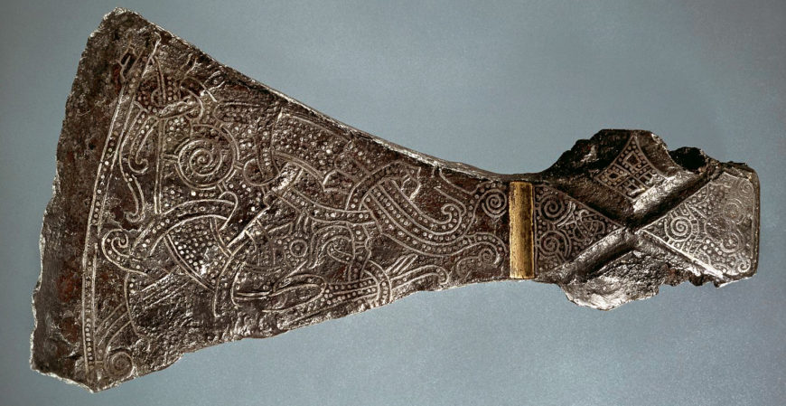 Photograph of an intricately engraved metal axe head featuring detailed swirling patterns and animal motifs. The axe head has a broken handle area with a visible gold band near the top, highlighting craftsmanship and decorative techniques.