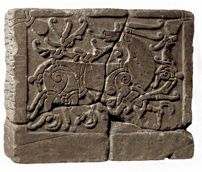 Photograph of an ancient carved stone relief depicting two stylized animals with spiral and linear patterns, possibly representing mythological or symbolic figures. The relief is divided into four broken pieces fitted together, showing detailed engravings and a border framing the scene.