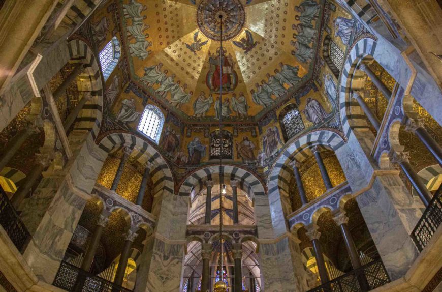 Photograph of an Palatine Chapel interior featuring a domed ceiling adorned with golden mosaics and religious figures. The structure includes striped arches, tall columns, and stained glass windows, highlighting intricate architectural and artistic details.