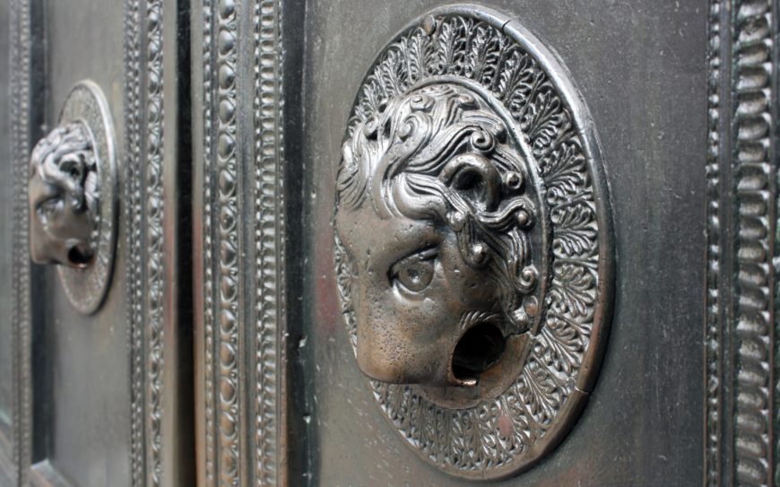 Photograph of an ornate metal door featuring a detailed lion head door knocker with an open mouth and intricate decorative border. The door has a textured surface with vertical panels and another smaller lion head knocker visible in the background.