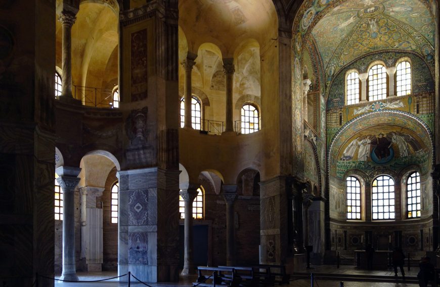 Photograph of a church interior showcasing Roman and Byzantine architectural elements with tall arches, ornate mosaics, and large arched windows allowing natural light. The space features detailed wall decorations, vaulted ceilings, and a prominent apse with intricate artwork and multiple window panes.
