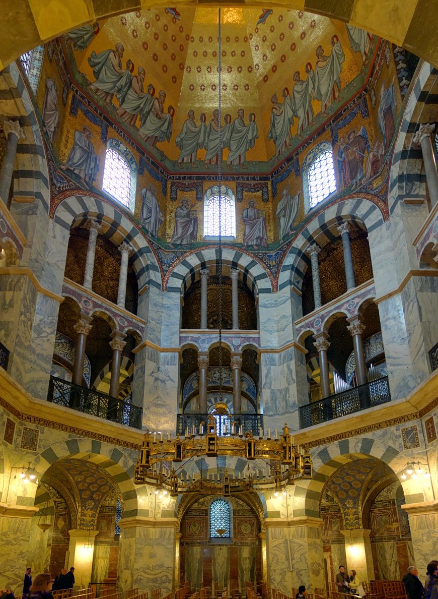 Photograph of an ornate cathedral interior showcasing Roman inspired architecture with striped marble columns, arched windows, and a large circular chandelier. Upper walls feature detailed mosaics of robed figures on a gold background, highlighting religious artistry and historical craftsmanship.