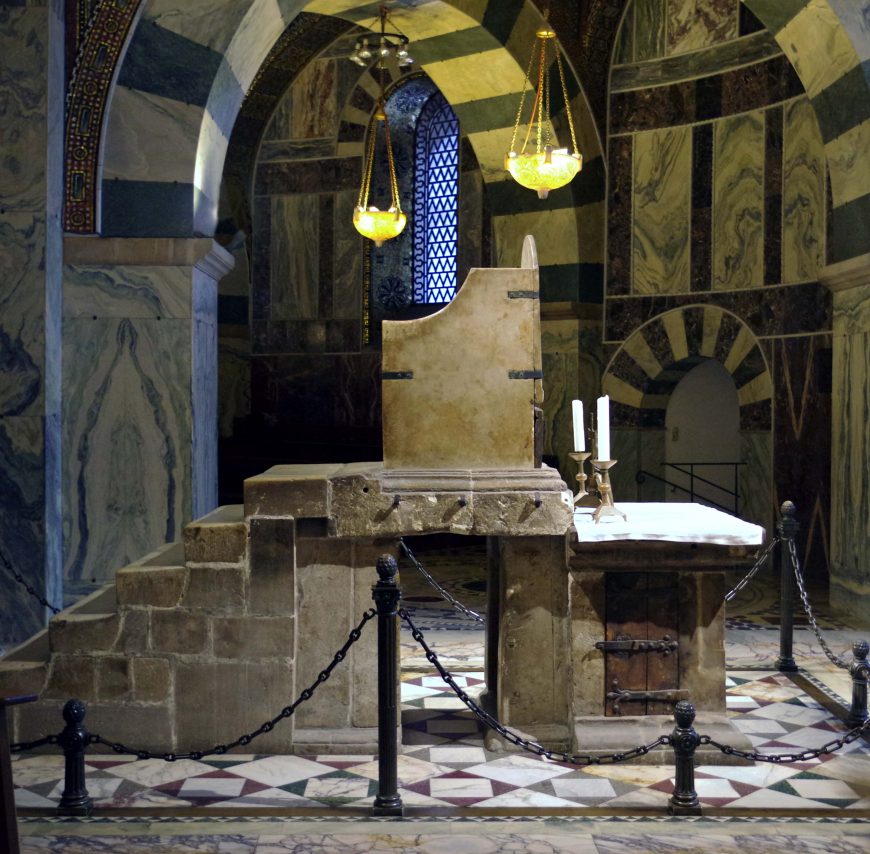 Photograph of a stone throne with a stepped platform and a small altar table with two candles, set inside a historic building with marble walls and arched ceilings. The area is cordoned off with black posts and chains, highlighting the throne's significance as a ceremonial or religious artifact.