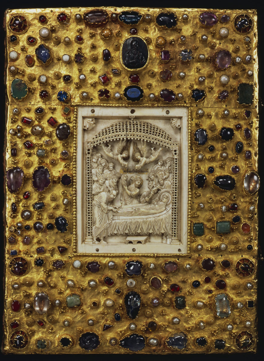 Photograph of an ornate medieval book cover featuring a central carved ivory panel depicting a religious scene surrounded by a gold background adorned with numerous colorful gemstones and pearls. The cover's intricate design highlights craftsmanship with a symmetrical arrangement of jewels in various shapes and sizes, emphasizing its historical and artistic significance.