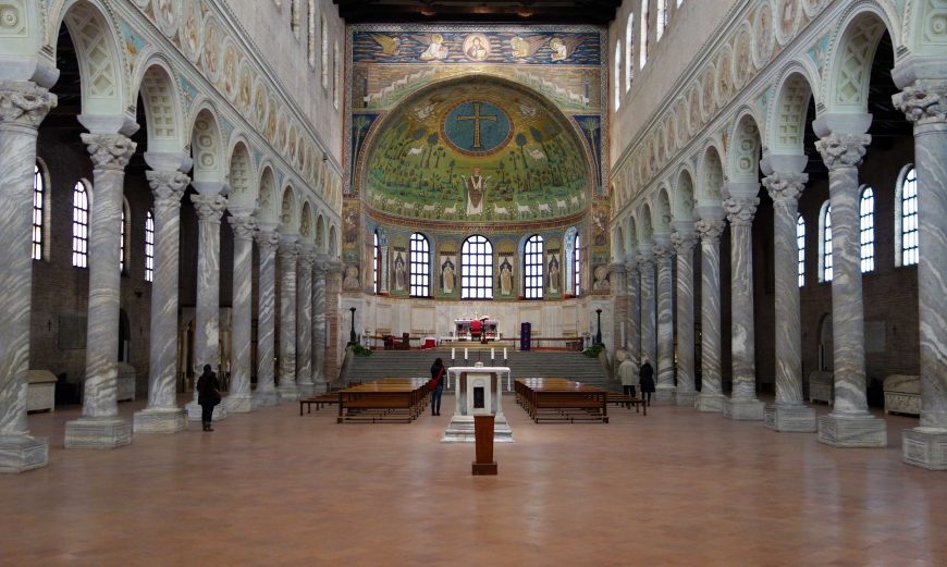 Photograph of a large, ornate church interior featuring tall marble columns on both sides and a decorated apse with religious mosaics and a large cross. The space includes wooden benches, high arched windows, and intricate architectural details emphasizing symmetry and grandeur.