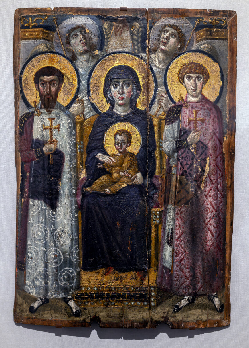 Religious icon painting depicting a central figure of the Virgin Mary holding baby Jesus, flanked by two saints with golden halos and ornate robes. The artwork features rich colors, intricate patterns, and symbolic elements such as crosses and halos, highlighting its spiritual significance.