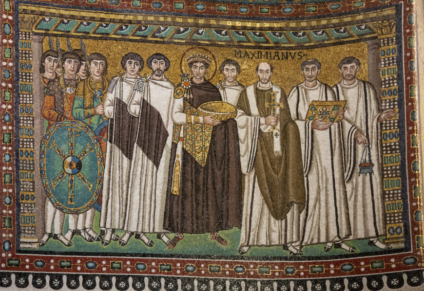 A Byzantine mosaic depicting Emperor Justinian I surrounded by clergy and soldiers, characterized by detailed robes, a golden halo, and a shield with a cross. The mosaic features vibrant colors, intricate patterns, and a Latin inscription above the bishop, highlighting religious and imperial authority.