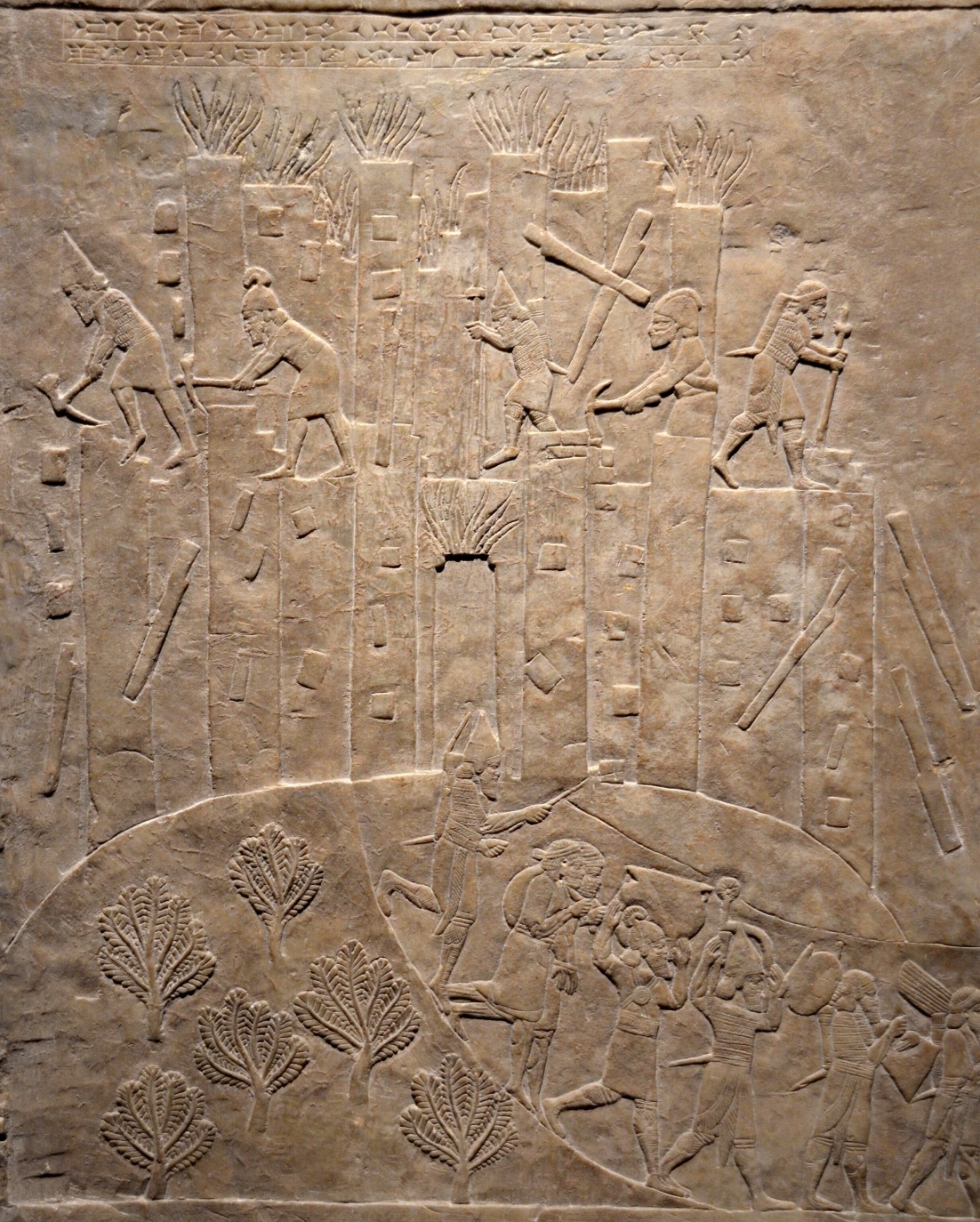 Sacking of Susa by Ashurbanipal bas-relief carving depicting ancient workers constructing a fortified wall with towers, using tools like hammers and chisels. The scene includes multiple figures on top of tall structures and others below, with stylized trees in the foreground.