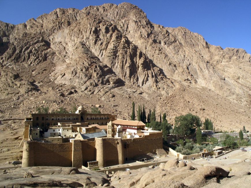 Photograph of a historic monastery complex situated at the base of a large, rugged mountain with clear blue sky. The structure features fortified stone walls, multiple buildings with domes and towers, surrounded by sparse greenery and desert terrain.