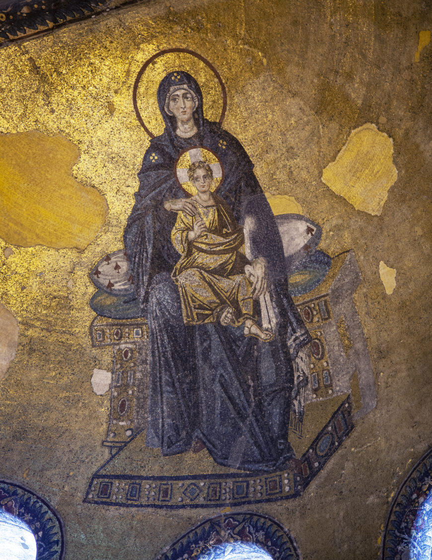 A mosaic artwork depicting a seated Madonna holding Child Jesus, both adorned with golden halos symbolizing holiness. The figures are set against a gold background with intricate patterns on the throne, highlighting Byzantine artistic style and religious iconography.