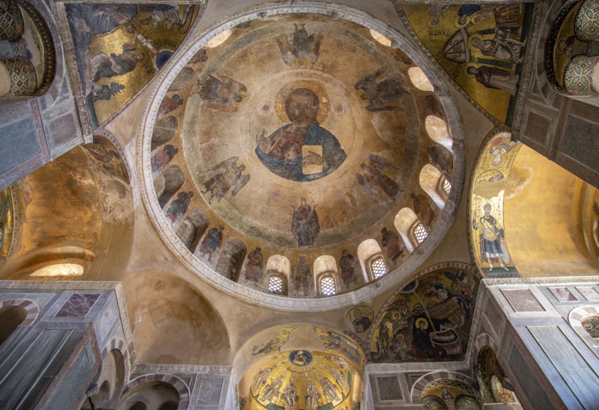 Photograph of an ornate church ceiling featuring a large central dome with religious frescoes and surrounding arches adorned with detailed iconography. Warm gold and earth tones highlight biblical scenes and figures, emphasizing historical and artistic significance of the interior architecture.