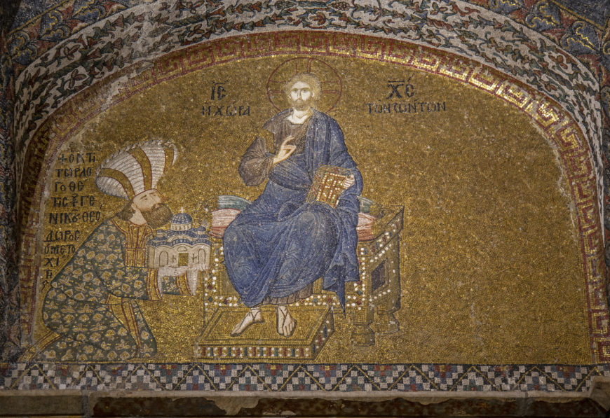 A Byzantine mosaic depicting a seated figure in blue robes holding a book, with a halo around the head, symbolizing a religious or divine person. To the left, a kneeling figure wearing a patterned robe and a tall headdress presents a model of a building, set against a gold background with Greek inscriptions above both figures.