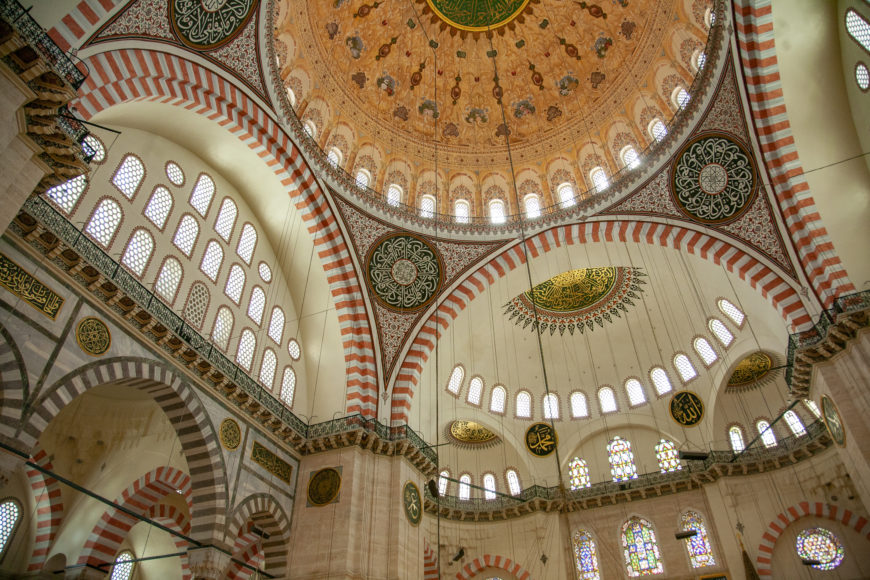 Photograph of an ornate mosque interior showcasing large domes with intricate geometric and floral patterns in gold, green, and red. Arched windows with stained glass and Arabic calligraphy medallions adorn walls, highlighting architectural and artistic details.