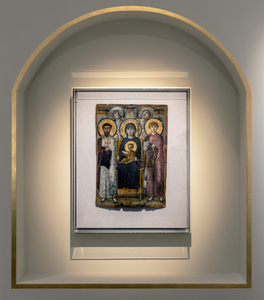 Photograph of a religious icon painting displayed within an arched, recessed wall niche with soft lighting. The artwork features five haloed figures, including a central seated figure holding a child, with detailed robes and a gold background.