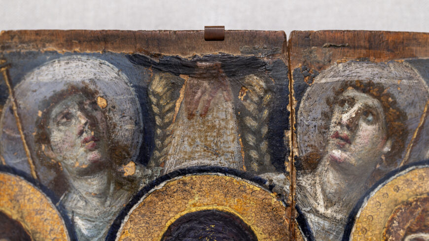 A close-up photograph of an ancient wooden icon panel featuring two angels with halos and a hand stretching down from the top, partially obscured by damage and wear. The panel shows faded paint, cracks, and darkened areas, highlighting its age.