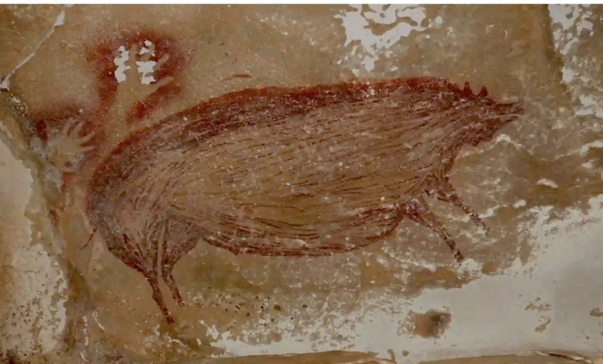 Photograph of prehistoric cave painting Warty pig depicting a large, brown, boar-like animal with detailed fur texture and a spiked back. Red handprints and abstract shapes surround the animal, indicating symbolic or ritualistic significance.