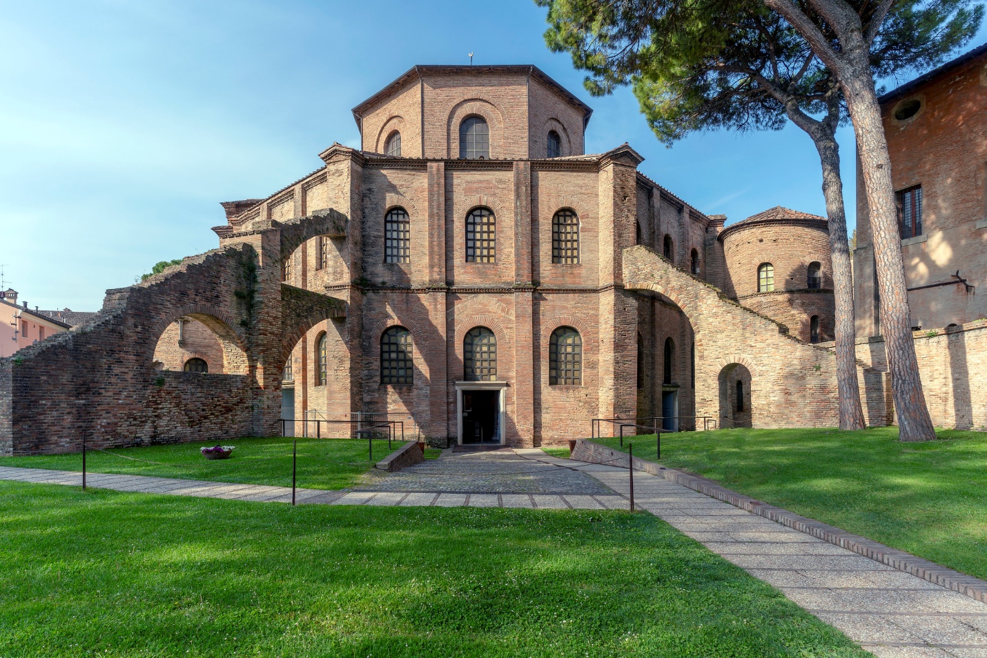 Photograph of an ancient brick building with arched windows and doorways, surrounded by green grass and trees under a clear blue sky. The structure features multiple levels and curved walls, highlighting historical architectural design and preservation.