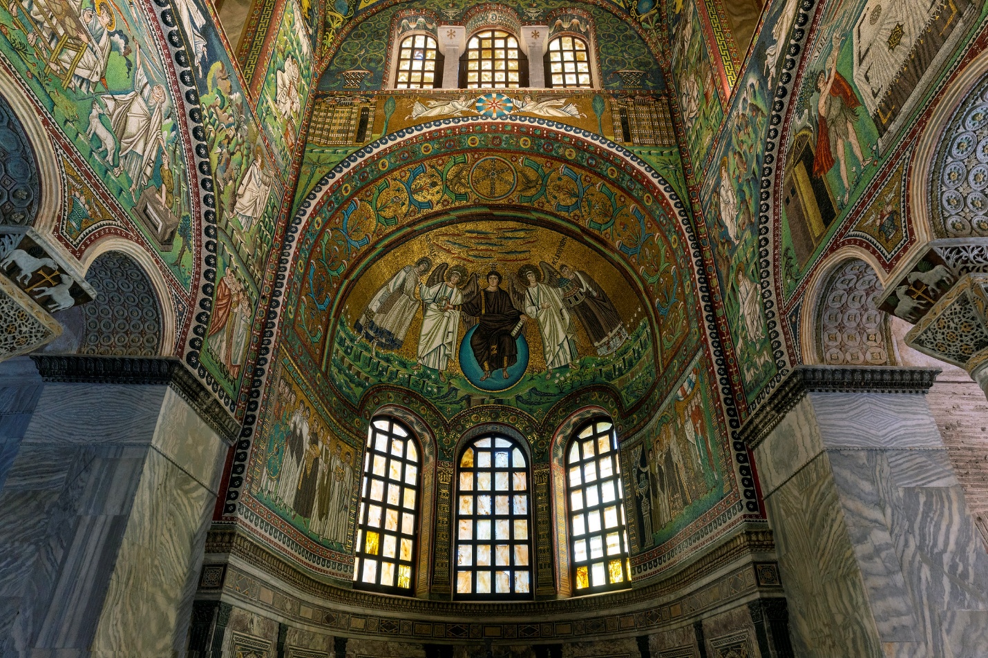 Photograph of an ornate church interior featuring detailed mosaics and stained glass windows. The central apse displays a prominent mosaic of a seated figure surrounded by angels, framed by intricate geometric and floral patterns in vibrant colors.