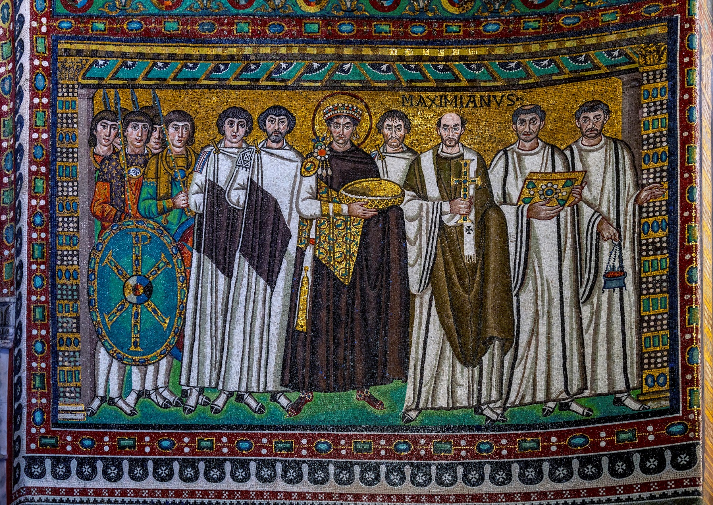 A Byzantine mosaic depicting Emperor Justinian I surrounded by clergy and soldiers, characterized by detailed robes, a golden halo, and a shield with a cross. The mosaic features vibrant colors, intricate patterns, and a Latin inscription above the bishop, highlighting religious and imperial authority.